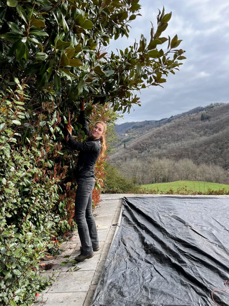 Deelgenote onderhoudt een groeiende voedselbos met struiken en plantgoed op het regeneratieve domein in de Vallée du Lot, Frankrijk, met heuvels op de achtergrond.